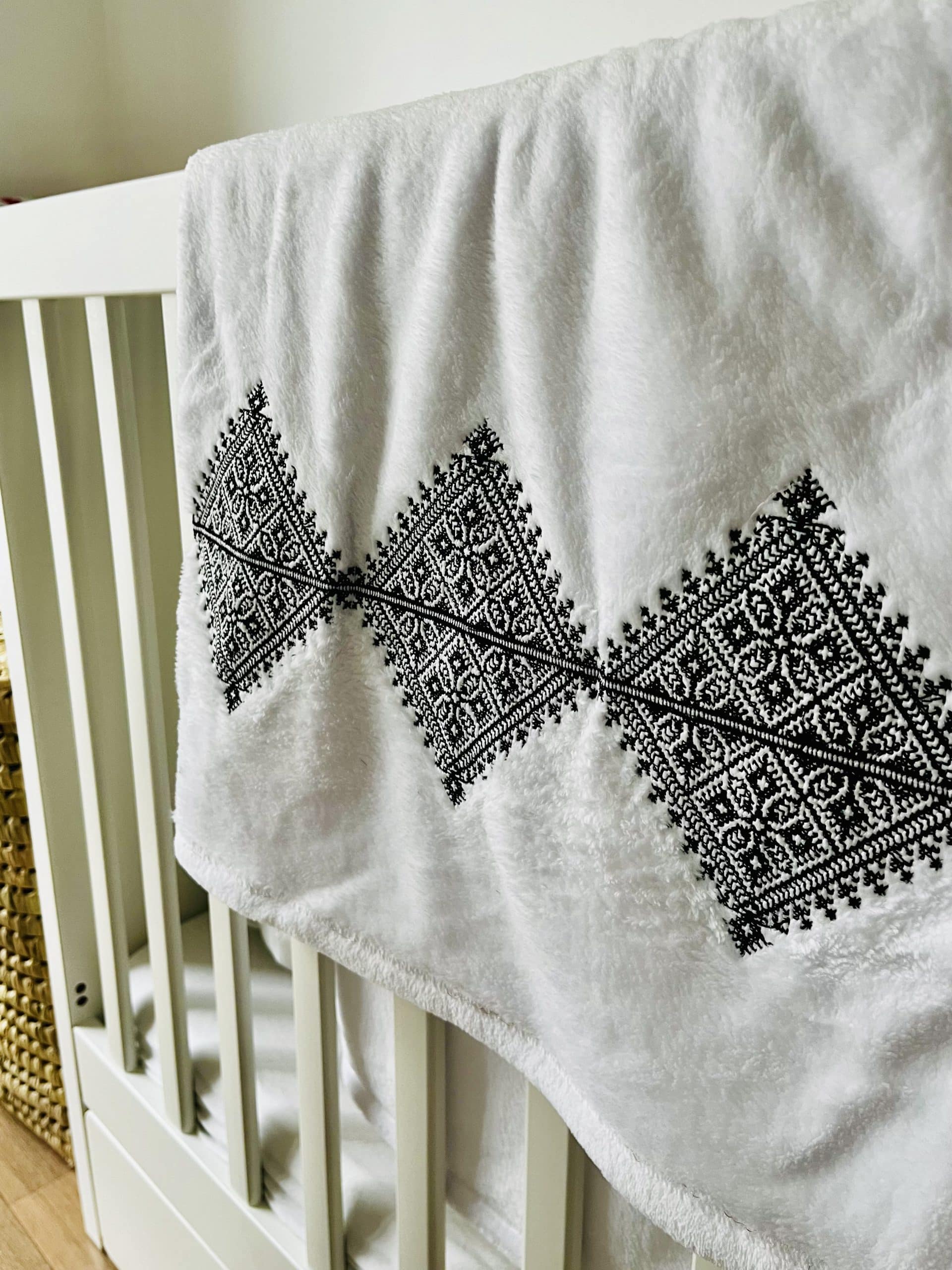 Plaid blanc avec broderie Fessi noir posé sur un lit bébé